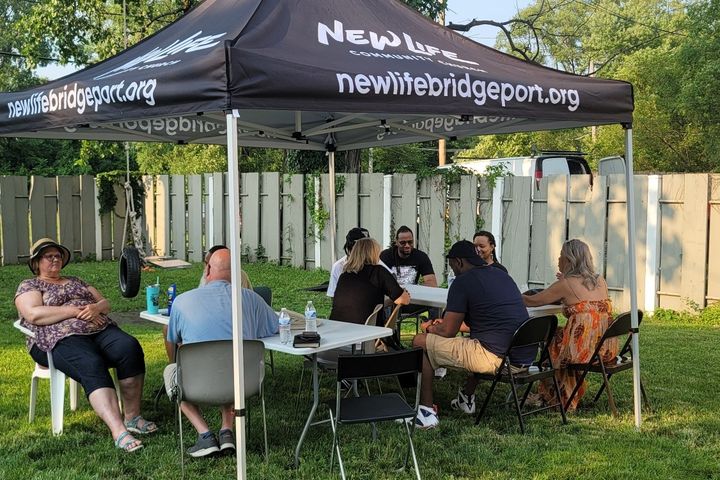 Neighbors gathered around tables under a tent for an outdoor Bible study